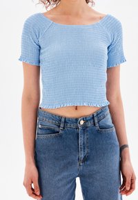 Modrá smockovaná krátká blůzka s raglánovými rukávy, zdobená volánkovým lemem. Nosená s džíny s vysokým pasem.