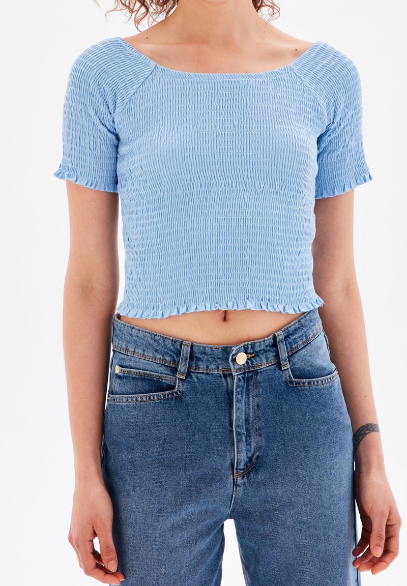 Modrá smockovaná krátká blůzka s raglánovými rukávy, zdobená volánkovým lemem. Nosená s džíny s vysokým pasem.