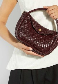 Woman holding a burgundy woven leather handbag with a curved handle and gold button clasp against a white textured top.