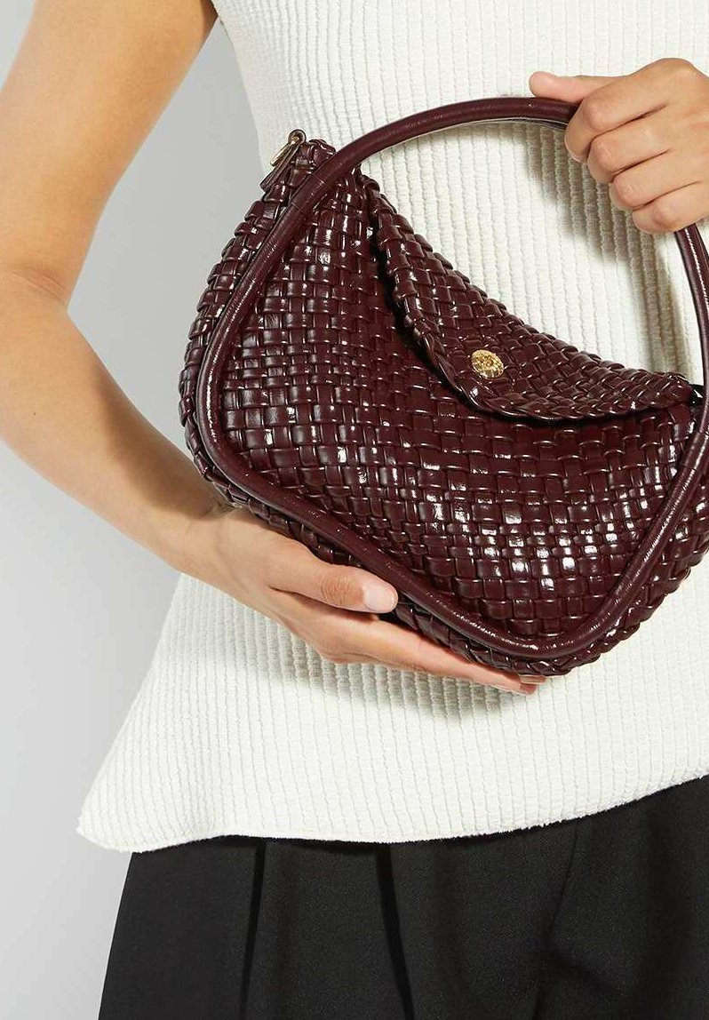 Woman holding a burgundy woven leather handbag with a curved handle and gold button clasp against a white textured top.