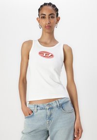 White sleeveless tank top featuring a red embroidered logo on the chest. Paired with light blue denim jeans. Smooth fabric texture.