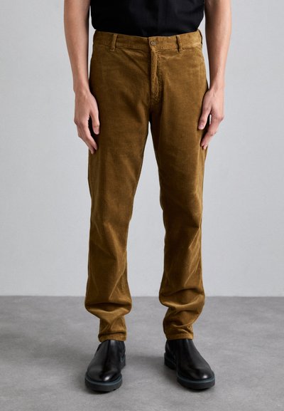 Pantaloni din catifea de culoare maro, cu croială dreaptă, cu o textură verticală vizibilă și un design clasic cu cinci buzunare, asortați cu cizme negre până la gleznă.