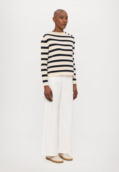 Maglione a righe blu navy e bianche con tre bottoni neri sulla spalla, abbinato a pantaloni bianchi a gamba larga e sneakers beige.