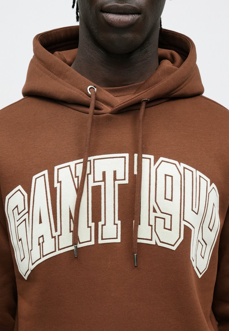 Brown hoodie made from soft material, featuring a large cream graphic text "GANT 1949" across the chest and silver hardware on the drawstrings.