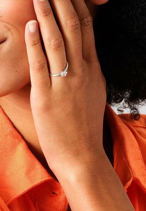 Silver ring with a small clear stone. Simple band design, worn on a hand with light brown skin, against an orange shirt.