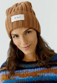 Un bonnet en tricot marron avec une texture côtelée et une étiquette blanche portant l'inscription "oui" en noir. Le design est doux et ajusté.