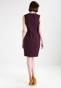 Žena s krátkými hnědými vlasy, oblečená v bezramínkových fialových šatech ke kolenům a hořčičně žlutých lodičkách, stojící zády k fotoaparátu na bílém pozadí.