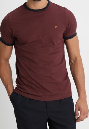 Mężczyzna ubrany w dopasowany bordowy t-shirt z czarnym kołnierzem i wykończeniami rękawów, zestawiony z czarnymi spodniami, stojący z jedną ręką w kieszeni.