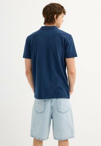 Camisa polo azul marino con cuello clásico, mangas cortas y dobladillo recto, combinada con pantalones cortos de mezclilla azul claro que tienen bolsillos traseros.