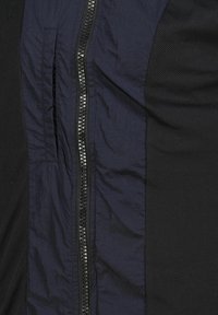 Giacca blu navy con zip lucida al centro, pannelli in rete texturizzati sui lati e finitura stropicciata.