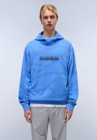 Hanorac albastru deschis cu un model texturat și logo frontal, având glugă cu șnur și manșete striate. Asociat cu pantaloni gri deschis.