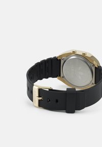 Fekete szilikonpántos óra arany színű házzal, nyolcszögletű formával és ezüst színű hátlappal. Fém csattal rendelkezik, amelyen logó részlet található.