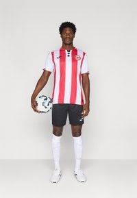 Camisa de futebol às riscas vermelhas e brancas com colarinho preto, calções pretos, meias brancas e uma bola de futebol. Inclui logotipo e detalhes da marca.