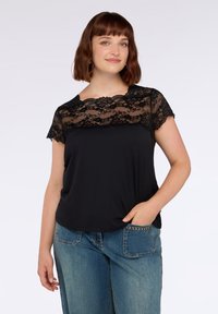 Giovane donna con capelli corti castani indossa un top nero con yoke in pizzo e maniche, abbinato a jeans blu, mentre sta in piedi contro uno sfondo semplice.