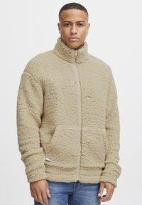 Beige fleece-jakke med høy krage, glidelås, elastiske mansjetter og doble frontlommer. Teksturen ser myk og fluffy ut.