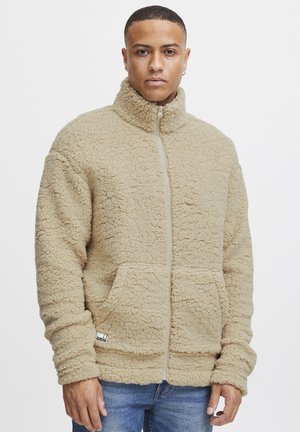 Tan fleece jacket with high collar, zip closure, elastic cuffs, and dual front pockets. Texture appears soft and fluffy.