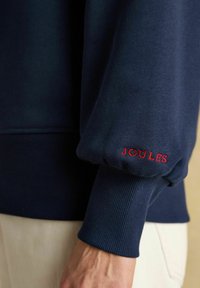 Jūras karaflīte sporta krekls ar elastīgām piedurknēm, ribotu tekstūru un sarkano "JOULES" logo labajā piedurknē. Neforšs dizains ar ērtu piegriezumu.