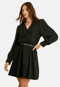 Rochie neagră cu un corp plisat, talie definită și mâneci lungi. Prezintă accente aurii pe umeri și un detaliu cu nasturi în față.