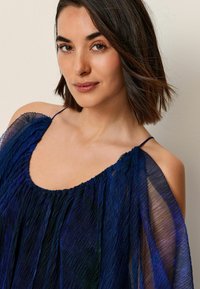 Rochie bleumarin cu bretele subțiri, material plisat și transparent, și decolteu adunat. Textură moale cu un design curgător și stratificat.