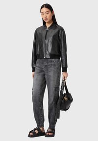 Schwarze Leder-Bomberjacke mit gerippten Bündchen, kombiniert mit grauen Cargohosen und schwarzen flachen Sandalen. Das Modell hält eine schwarze Tasche.