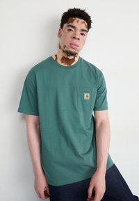 Green cotton t-shirt with a crew neck and short sleeves. Features a small chest pocket and a logo patch. Worn with dark denim jeans.
