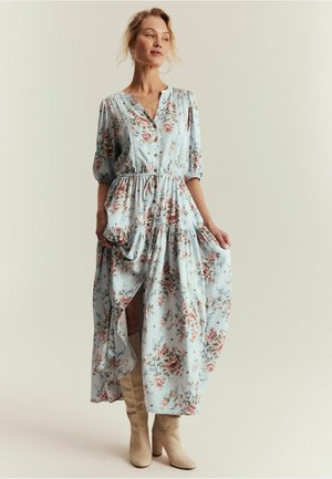 Kvinde iført en lyseblå blomsterprintet midikjole med knapper og trekvart ærmer, kombineret med beige støvler med hæl, stående mod en ensfarvet baggrund.