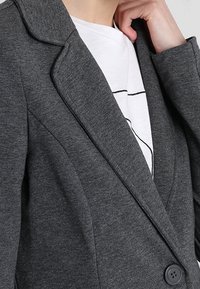 Šedý blazer vyrobený z hladké tkaniny, s klopovým límcem, zapínáním na jeden knoflík a přiléhavým střihem.
