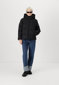 Geacă puffer neagră cu glugă, fermoar frontal și textură matlasată, asortată cu blugi albaștri cu un model în carouri la mansete. Încălț�ăminte neagră.