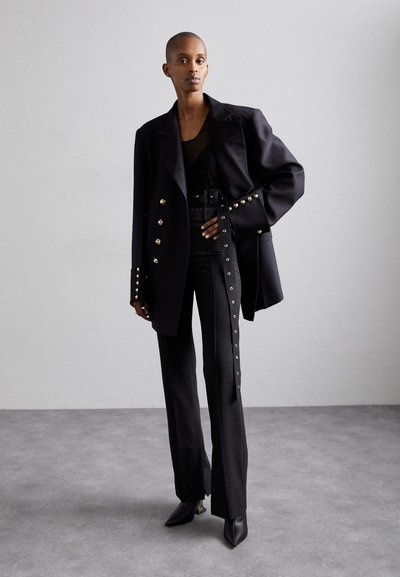 Black tailored blazer with gold button accents, fitted black top, high-waisted trousers with side detailing, and pointed black shoes.