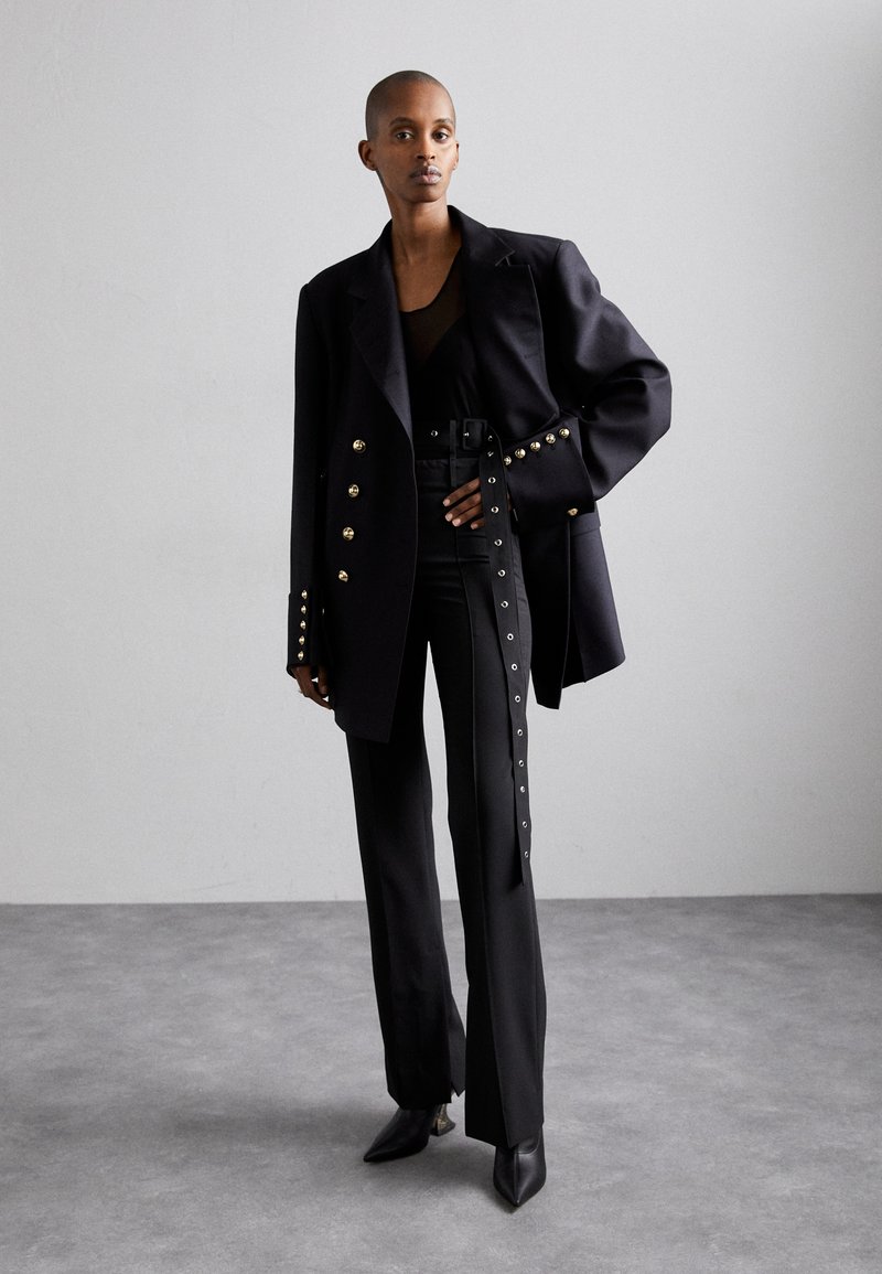 Blazer noir ajusté avec des accents de boutons dorés, haut noir cintré, pantalon taille haute avec des détails sur les côtés, et chaussures noires à bout pointu.