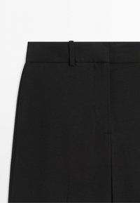 Close-up van het bovenste gedeelte van een zwarte broek met tailleband en een riemlus op een witte achtergrond.