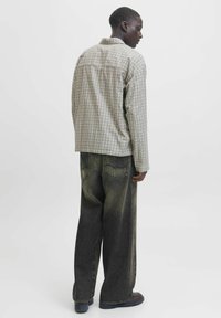 Kariertes Hemd in Beige und Braun, geknöpft, mit einem lässigen Schnitt. Kombiniert mit lockeren, dunkelgrauen Jeans und dunklem Schuhwerk. Rückansicht.