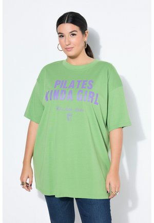 Mujer con el pelo oscuro recogido, lleva pendientes de aro dorados y una camiseta verde holgada con el texto "PILATES KINDA GIRL" en color púrpura.