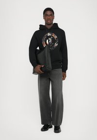Sudadera negra con un diseño circular impreso, pantalones vaqueros anchos grises y zapatos negros. Sosteniendo un bolso de cuero negro, contra un fondo blanco liso.