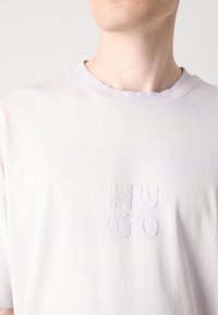 Lys grå, kortærmet T-shirt lavet af bomuld, med hævet hvid broderet tekst "HUGO" i midten. Rund krave.