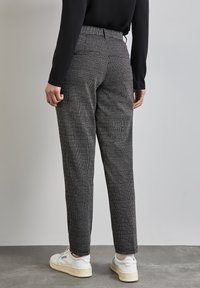 Pantaloni a scacchi bianco e nero, design a vita alta, con una vestibilità sartoriale e parte anteriore piatta. Abbinati a sneakers bianche su un pavimento grigio.