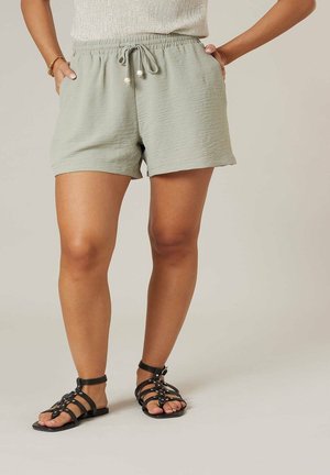 Femme portant un short vert clair à cordon de serrage et des sandales plates noires à lanières, debout les mains dans les poches.