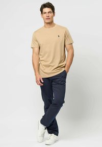 Egy férfi bézs pólót, tengerészkék nadrágot és fehér sportcipőt visel, az egyik kezét zsebébe dugva, egy sima háttér előtt áll.