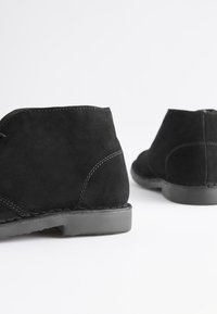 Black suede ankle boots with a low rubber sole, featuring a smooth texture and subtle stitching details on the sides.