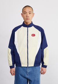 Marinblå och gräddfärgad vindjacka med vita detaljer, hög krage och framdragkedja. Har en röd Nike Air-logotyp på bröstet.