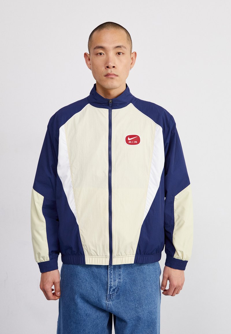 Marinblå och gräddfärgad vindjacka med vita detaljer, hög krage och framdragkedja. Har en röd Nike Air-logotyp på bröstet.