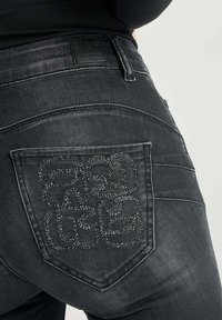 Des jeans en denim noir présentent une poche arrière texturée avec un design de couture complexe. Le tissu a une finition lisse et soyeuse avec une légère usure.