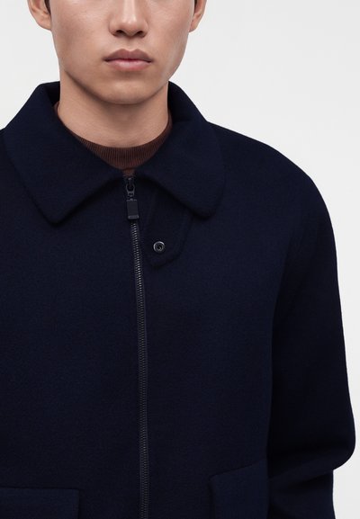 Jeune homme portant un manteau bleu marine foncé avec col et fermeture éclair devant, par-dessus un pull col rond marron, sur un fond clair uni.