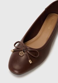 Ballerine en cuir marron avec bout arrondi, ornée d'un nœud et d'un charm doré à l'avant, surface lisse et semelle intérieure neutre.