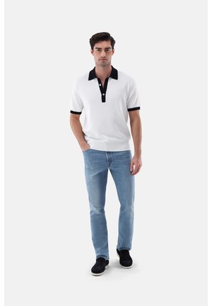 Mann steht und trägt ein weißes kurzärmeliges Polo mit schwarzem Kragen und Manschetten, hellblaue Jeans und schwarze Slipper auf weißem Hintergrund.