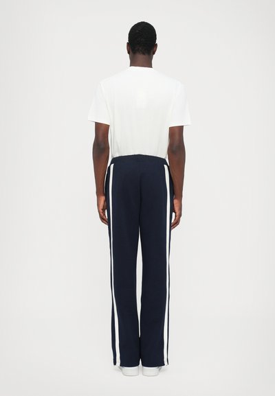 Navy track pants with white side stripes, soft fabric, straight legs, elastic waistband, paired with a plain white t-shirt. Back view.
