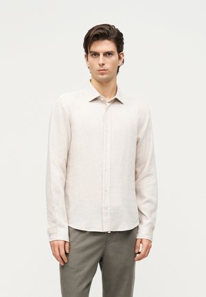 Mladý muž s krátkymi hnedými vlasmi, ktorý má na sebe svetlobéžovú košeľu s dlhým rukávom na gombíky a sivé nohavice, stojí pred jednoduchým pozadím.