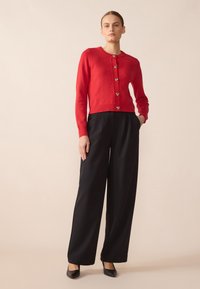 Cardigan rouge avec encolure ronde, fermeture à boutons sur le devant et ourlet côtelé. Associé à un pantalon large noir et des talons noirs.