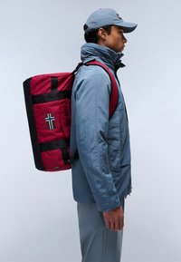 Un sac à dos rouge et noir avec un patch du drapeau norvégien, présentant des accents de logo et des bretelles épaisses, porté sur une veste bleue vue de profil.