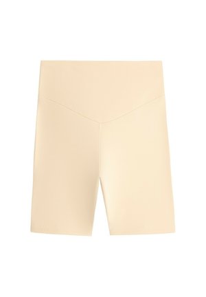 Femme debout portant un t-shirt à manches longues rayé, un short de vélo beige, des chaussettes blanches, des baskets et un sac bandoulière sur un fond gris.